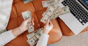 Side Hustles And Businesses To Make Money ASAP Two hands holding money beside a laptop, representing online side hustles and businesses to make money ASAP.