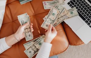 Side Hustles And Businesses To Make Money ASAP Two hands holding money beside a laptop, representing online side hustles and businesses to make money ASAP.