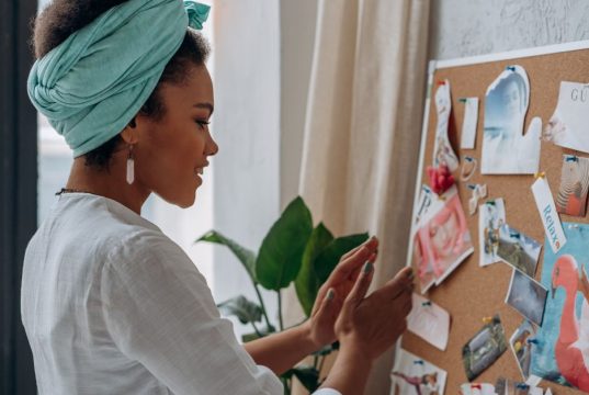 Creating Your Vision Board: Turn Your Dreams into Reality A Black woman smiling while looking at her vision board filled with photos and quotes, symbolizing creativity and purpose in creating your vision board.