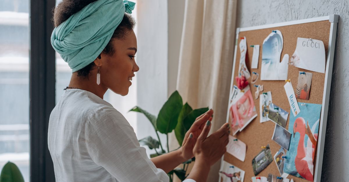 A Black woman smiling while looking at her vision board filled with photos and quotes, symbolizing creativity and purpose in creating your vision board.