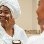 A Black woman smiling at her reflection while applying a white cream to her cheek, showing the results of using the best skincare products for glowing, healthy skin.
