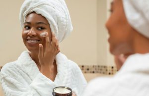 The Best Skincare Products For Every Skin Type A Black woman smiling at her reflection while applying a white cream to her cheek, showing the results of using the best skincare products for glowing, healthy skin.