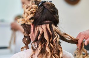 How To Style Your Hair Like A Celebrity A woman with blonde and brown wavy hair facing away, with a stylist’s hands arranging her hair, demonstrating how to style your hair like a celebrity.
