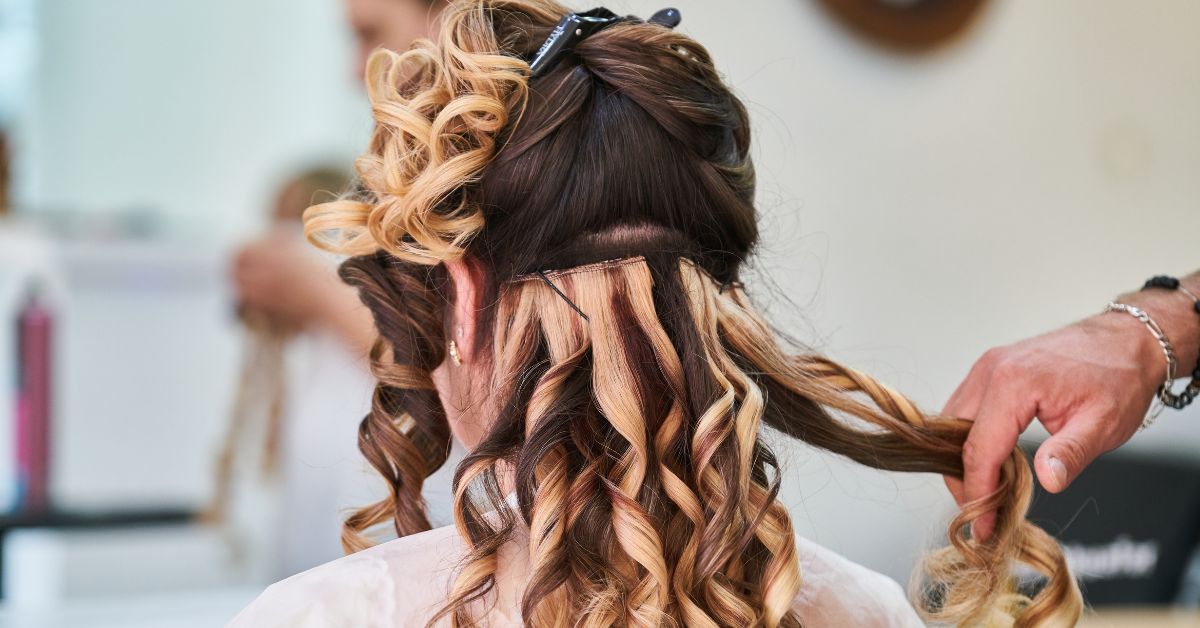 A woman with blonde and brown wavy hair facing away, with a stylist’s hands arranging her hair, demonstrating how to style your hair like a celebrity.