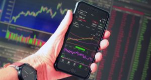 The Top 10 Stocks To Invest In For 2024 – 2025 A hand holding a smartphone displaying an AAPL stock market chart with green and red trading indicators, representing the top stocks to invest in 2024–2025.