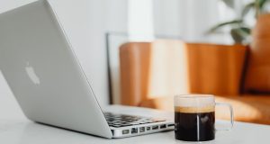 Morning Motivation Tips to Boost Your Energy and Productivity Morning motivation workspace setup showing an Apple laptop and coffee cup on a bright table, with sunlight streaming through the window and a blurred couch and green plant in the background — perfect for morning motivation tips to boost your energy and productivity.