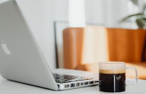 Morning Motivation Tips to Boost Your Energy and Productivity Morning motivation workspace setup showing an Apple laptop and coffee cup on a bright table, with sunlight streaming through the window and a blurred couch and green plant in the background — perfect for morning motivation tips to boost your energy and productivity.