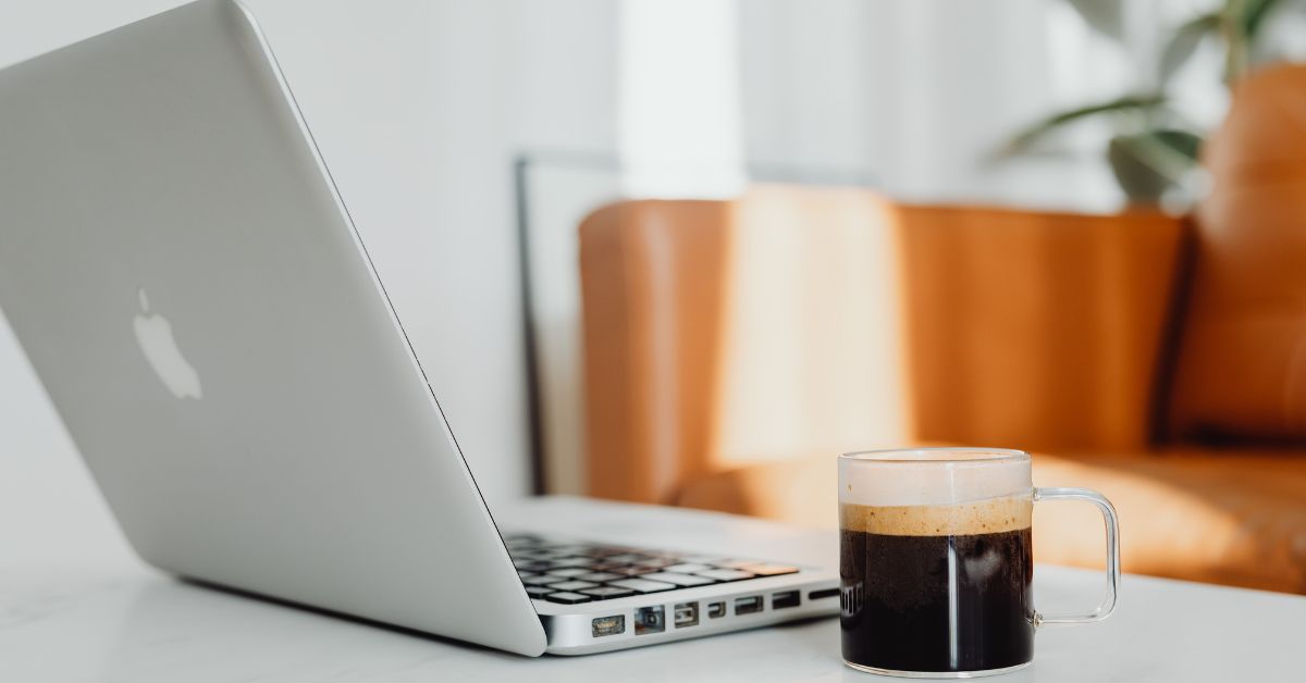Morning motivation workspace setup showing an Apple laptop and coffee cup on a bright table, with sunlight streaming through the window and a blurred couch and green plant in the background — perfect for morning motivation tips to boost your energy and productivity.