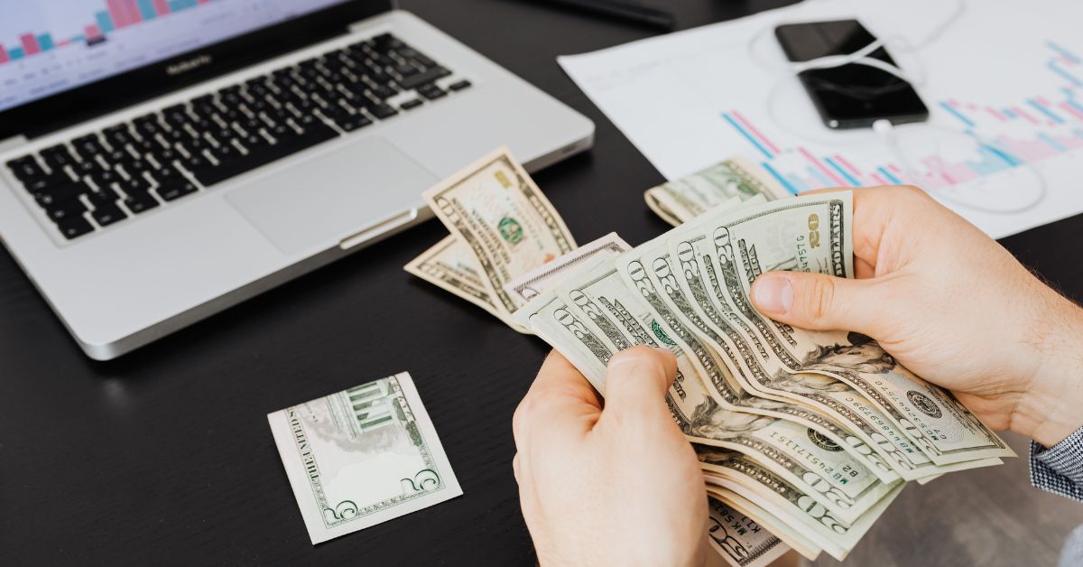 Hands counting money with laptop, document, and phone in the background, illustrating how to get funding for your business and manage finances effectively.