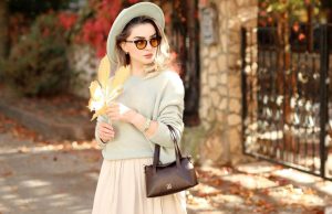 How To Look Rich And Classy On A Budget A stylish woman wearing a neutral outfit, hat, and sunglasses during autumn, holding a brown leaf with sunlight on her — showing how to look rich and classy on a budget.