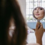 Woman looking at her reflection in a small mirror and smiling softly, representing self-acceptance and confidence — a visual for simple ways to overcome self-doubt.