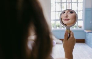 8 Simple Ways To Overcome Self-Doubt Woman looking at her reflection in a small mirror and smiling softly, representing self-acceptance and confidence — a visual for simple ways to overcome self-doubt.
