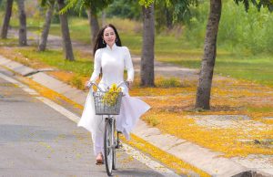 New Beginnings: A Fresh Start to Your Best Chapter Yet! Woman in white riding a bicycle on a road beside a tree-lined pavement covered with green grass and orange leaves, smiling softly and embracing new beginnings.