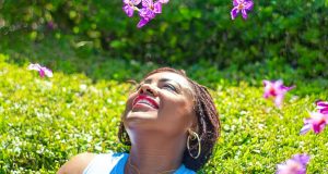 How To Stay Motivated When You’re Feeling Down A woman lying on green grass surrounded by pink flower petals, smiling peacefully under bright sunlight, representing positivity, peace, and how to stay motivated when you’re feeling down.