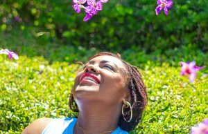 How To Stay Motivated When You’re Feeling Down A woman lying on green grass surrounded by pink flower petals, smiling peacefully under bright sunlight, representing positivity, peace, and how to stay motivated when you’re feeling down.