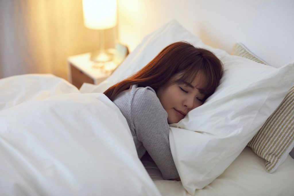 A woman sleeping peacefully on a white pillow and bed, wearing a grey top, showing calm and relaxation as part of daily rituals to sleep better.