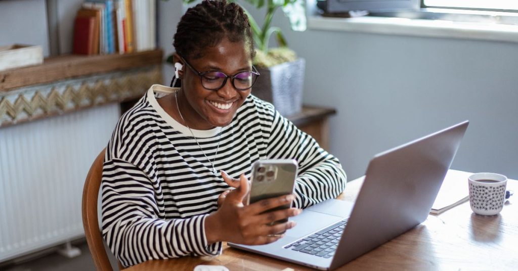 A smiling black woman working remotely on a laptop, checking her phone for dollar payments — how to get paid in dollars from Nigeria.