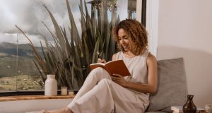 10 Simple Self-Care Routines Every Busy Woman Can Start Today A woman enjoying a peaceful morning self-care moment, sitting by the window with a journal and pen, practicing simple self-care routines for busy women.