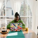African woman smiling while working on a laptop at a home office table with coffee, sunlight and white curtains, illustrating smart ways to diversify your income in Nigeria