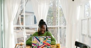 Smart Ways to Diversify Your Income in Nigeria (Even with a 9-5 Job) African woman smiling while working on a laptop at a home office table with coffee, sunlight and white curtains, illustrating smart ways to diversify your income in Nigeria