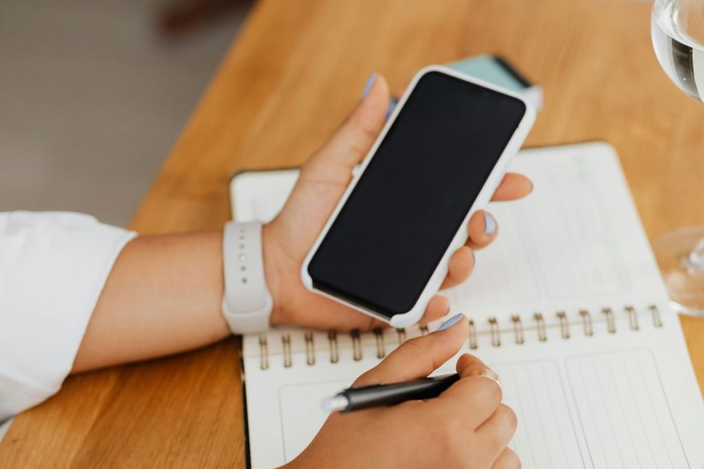 Hand holding a smartphone above a notebook, showing how to check Infinix or Tecno phone warranty in Nigeria