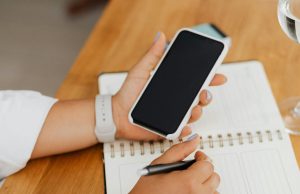 How to Check If Your Infinix or Tecno Phone Warranty Still Applies in Nigeria Hand holding a smartphone above a notebook, showing how to check Infinix or Tecno phone warranty in Nigeria