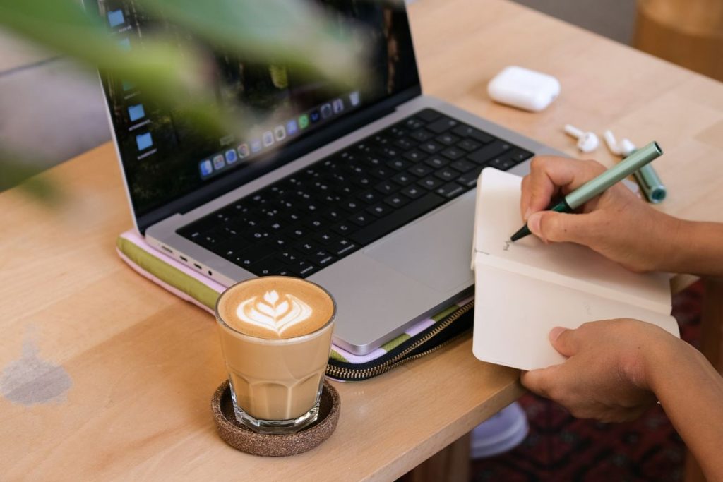 Hands writing in a notebook beside a laptop and coffee, showing how to set realistic goals and actually achieve them